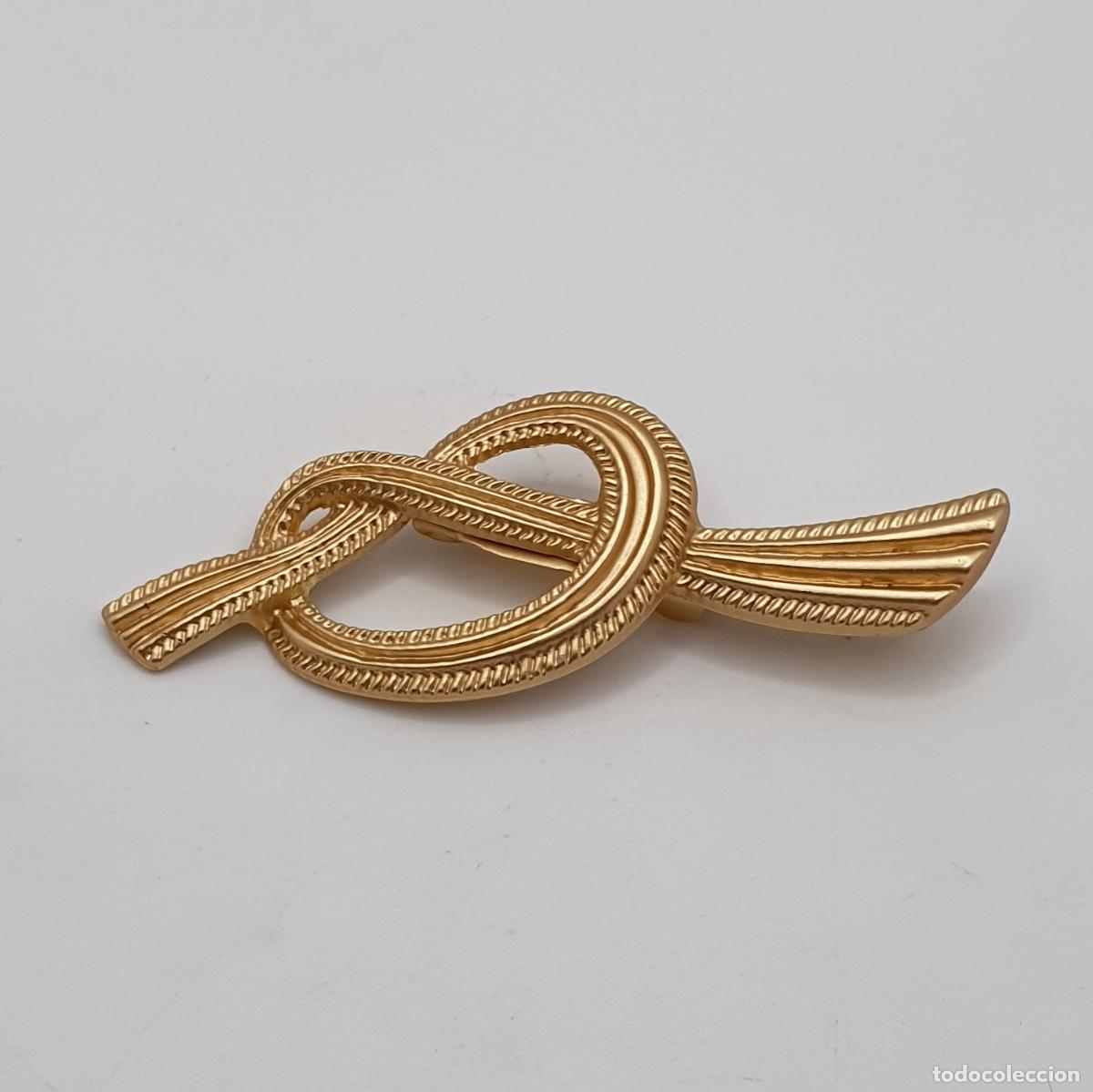 Joyeria: Elegante y sofisticado broche de cinta formando un nudo con acabados en oro mate.