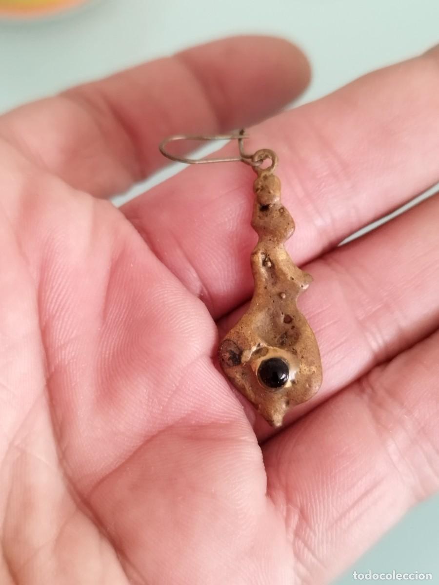 Joyeria: Antiguo pendiente artesanal o arqueol&oacute;gico, posiblemente hecho de metal con piedra oscura central.