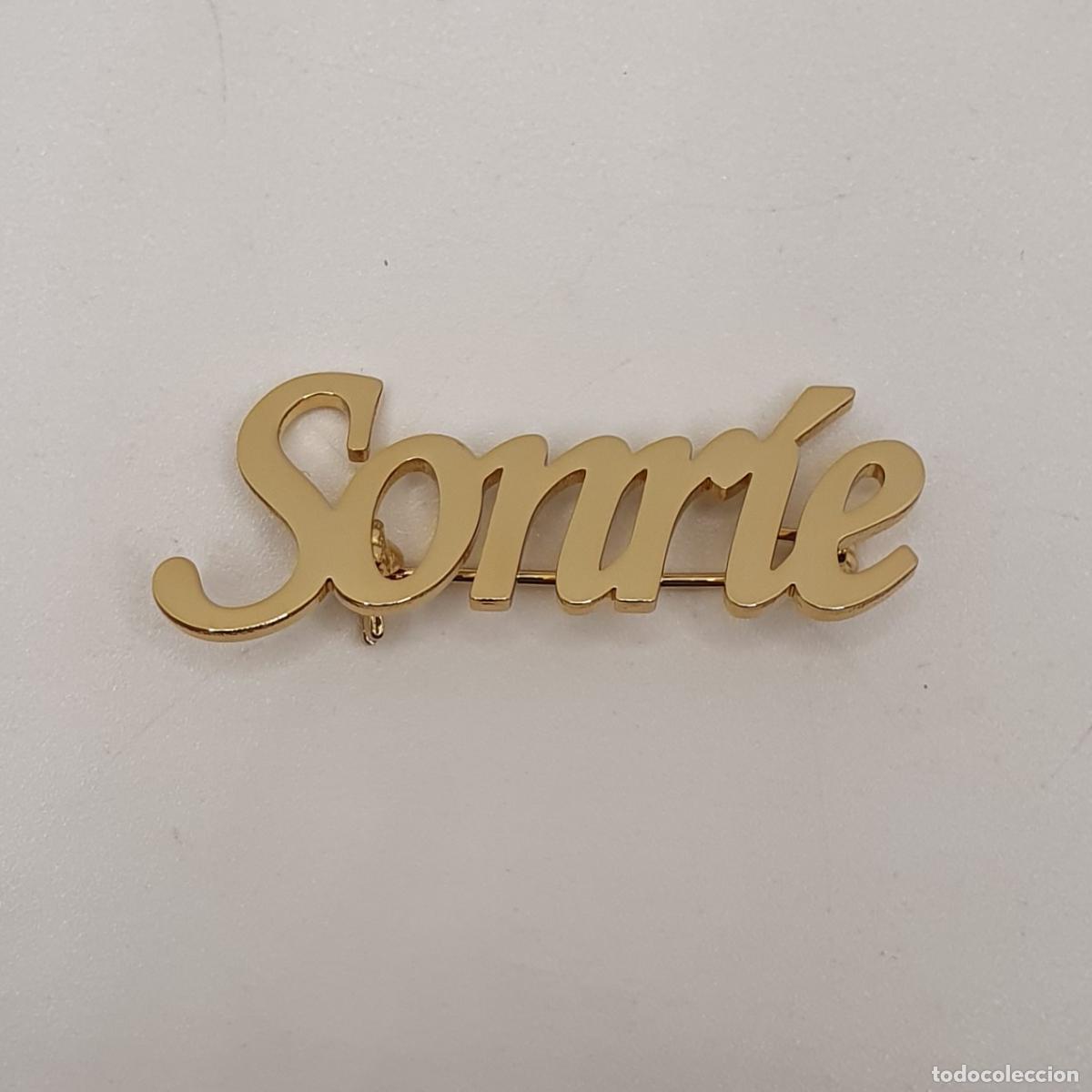 Joyeria: Exquisito broche con acabados en oro con la palabra SONR&Iacute;E para robar sonrisas all&aacute; donde vayas.