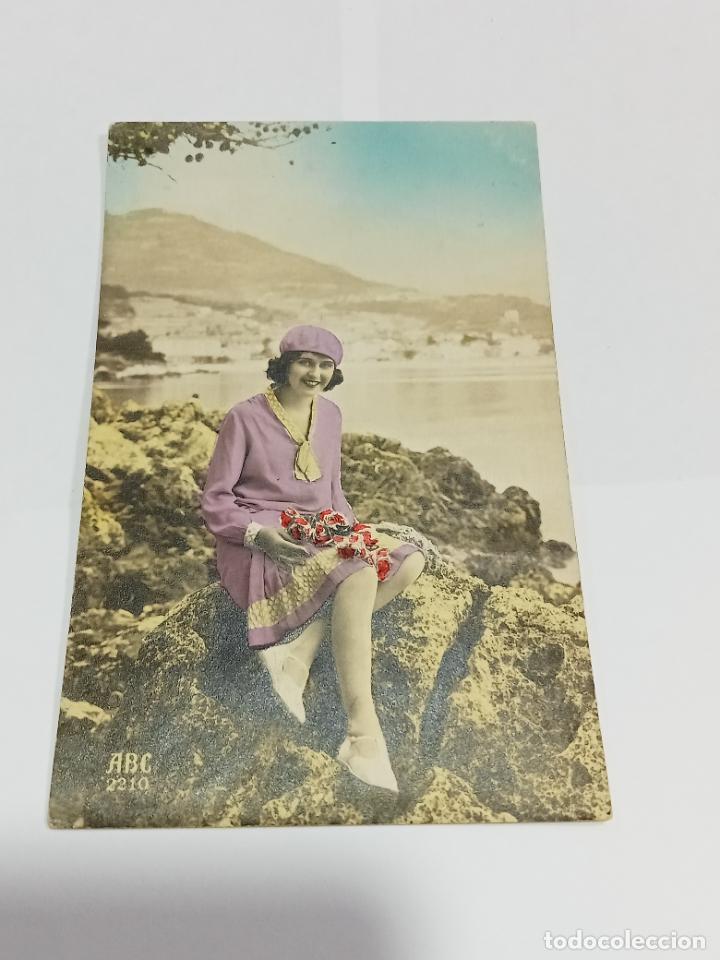 Cartoline: TARJETA POSTAL DE MUJERES. MUJER SENTADA ENTRE ROCAS CON VESTIDO MORADO Y RAMO DE FLORES