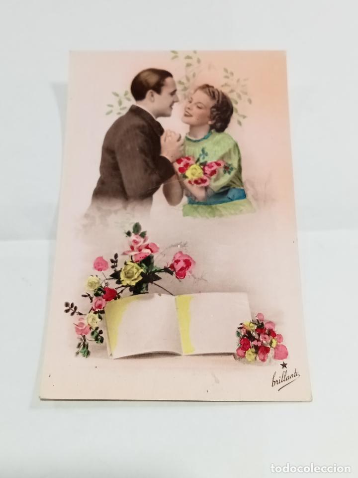Postais: TARJETA POSTAL. PAREJA DE ENAMORADOS MIR&Aacute;NDOSE RODEADOS DE FLORES Y CON LIBRO POR ESCRIBIR